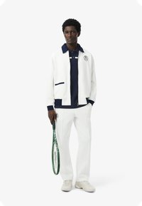 Chaquetón blanco con cuello y detalles en azul marino, combinado con una polo azul marino y pantalones blancos. Sosteniendo una raqueta de tenis verde.