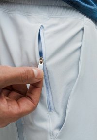 Lyseblå træningsbukser har en lynlåslomme. Lynlåsens tr�ækstang er sølvfarvet med en blå accent. Stoffet er glat og strækbart.
