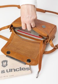 Braune Leder-Crossbody-Tasche mit weicher Textur, Druckverschluss und gestreiftem Innenfutter. Enthält eine rote Reißverschlusstasche und einen markierten Stoffbeutel.