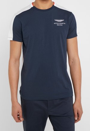 Mann trägt ein marineblaues Aston Martin Racing T-Shirt mit weißen Schultern und dunkler Hose, steht vor einem einfarbigen Hintergrund.