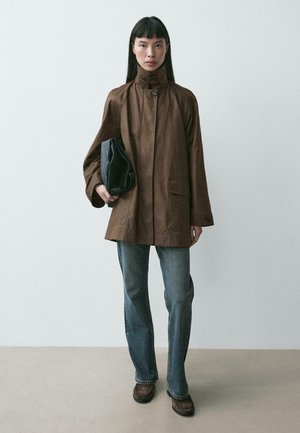 Giovane donna con capelli lisci e scuri indossa una giacca marrone con bottoni, jeans blu e scarpe marroni, tenendo un ombrello nero pieghevole.