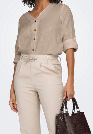 Femme portant un chemisier beige boutonné avec manches retroussées et un pantalon taille haute assorti, tenant un sac à main texturé marron foncé dans la main droite.