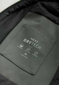 Grøn stofmærkat på indersiden af en jakke med "NEXT DRYTECH" branding, ikoner for vandtæt, tapede sømme, letvægts og størrelse "M."