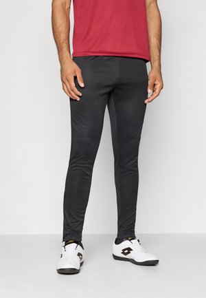 Sorte treningbukser laget av glatt, elastisk materiale, med en slim fit-design og glidelåslommer, sammen med hvite joggesko.