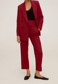 Persoon in een op maat gemaakte donkerrode dubbelgeknoopte blazer en bijpassende cropped broek, met zwarte instappers en een zwart top.