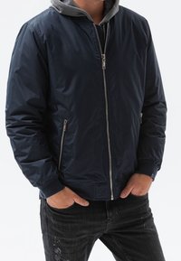 Marinblå bombarjacka med grå huva, dragkedja, sidfickor, ribbade muddar och slät, glänsande yta.