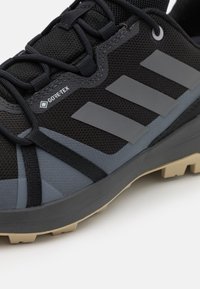 Černá atletická obuv se šedými akcenty, s textilním horním materiálem, texturovanou gumovou podrážkou a brandingem GORE-TEX. Zajímavý design se třemi pruhy.