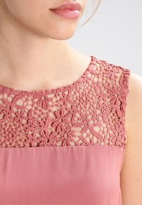 Robe rose avec un col en dentelle florale et un corps en tissu uni et lisse. Le design en dentelle inclut des motifs ajourés et des détails texturés.