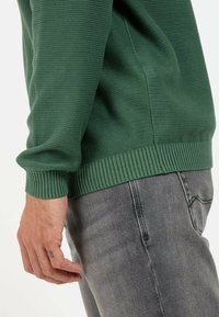 Green ribbed knit sweater with textured pattern and cuffed sleeves, paired with grey denim pants, partially visible hand on side.