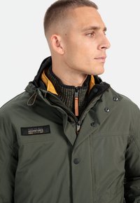 Olivengrøn jakke med lynlåslukning, trykknapper og en aftagelig hætte. Foret med gul stof og har en struktureret strikkrave.