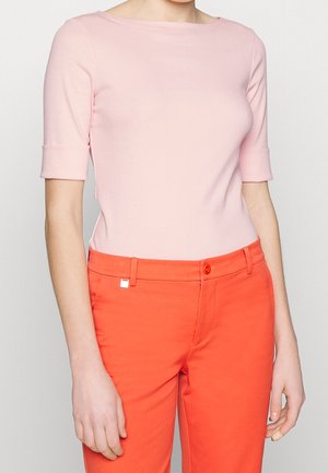 Mujer con una blusa de manga hasta el codo de color rosa pastel y pantalones de talle alto de color naranja brillante, de pie sobre un fondo blanco.