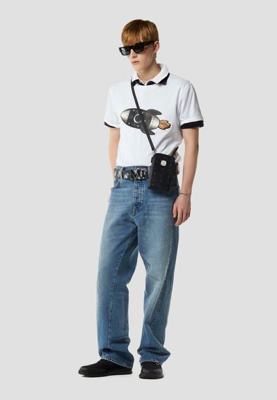 Jonge man met zwarte zonnebril, wit poloshirt met raketprint, losse blauwe jeans, zwarte sneakers, een riem en een kleine zwarte schoudertas.