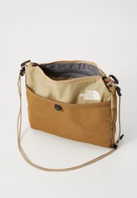 Lys brun crossbody-veske laget av slitesterkt stoff, med en mesh-frontlomme, en glidelåslukking og justerbar stropp. Hvit logo detalj til stede.