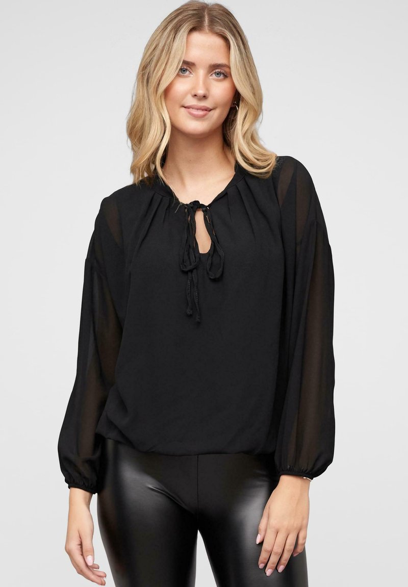 Blusa leggera nera con scollo rotondo, dettaglio con fiocco sul davanti e maniche lunghe trasparenti. Abbinata a leggings neri lucidi.