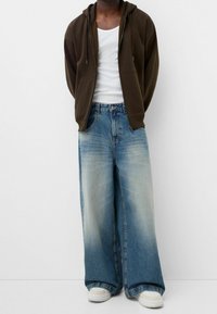 Felpa zip-up marrone scuro, canotta bianca, jeans a gamba larga blu chiaro con una texture sbiadita e sneakers bianche. I jeans presentano il classico design a cinque tasche.