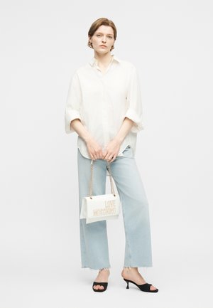 Γυναίκα που φοράει λευκό πουκάμισο σε μεγάλο μέγεθος και ανοιχτόχρωμο τζιν, κρατώντας λευκή τσάντα Love Moschino με χρυσές αλυσίδες ως χερούλια, φορώντας μαύρα πέδιλα με ανοιχτά δάχτυλα.