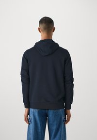 Hanorac cu glugă de culoare albastru marin, cu o textură netedă și manșete ribbate; purtat cu jeanși cargo de culoare albastru deschis, cu buzunare și o croială lejeră.