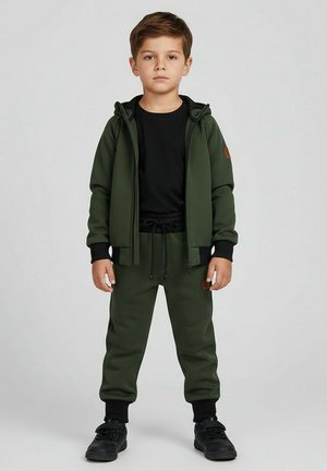 Chłopiec stojący, ubrany w ciemnozieloną kurtkę z kapturem i pasujące spodnie, czarną koszulkę i czarne trampki, neutralne tło.