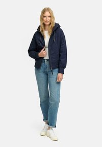 Giacca bomber leggera blu navy con cappuccio, polsini elasticizzati e chiusura con zip. Indossata con una maglietta bianca e jeans azzurri chiari.