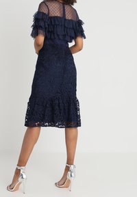 Robe en dentelle bleu marine avec manches transparentes à volants en couches, corsage ajusté et ourlet inférieur évasé. Talons à bride argentés à la cheville complètent le look.