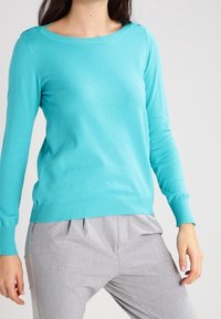 Turquoise long-sleeve sweater made of soft material, features ribbed neckline and hem, paired with light gray pleated pants.
