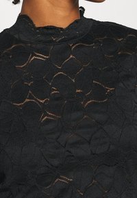 Sort langærmet bluse med indviklede blonder og blomster mønstre, tekstureret finish og høj hals.