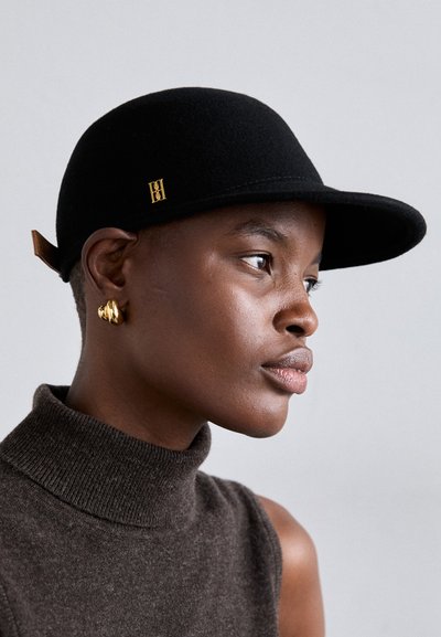 Gorra de fieltro negro con visera curva, con un detalle de logotipo dorado y una correa de cuero en la parte posterior. Modelo vistiendo un suéter de cuello alto marrón.