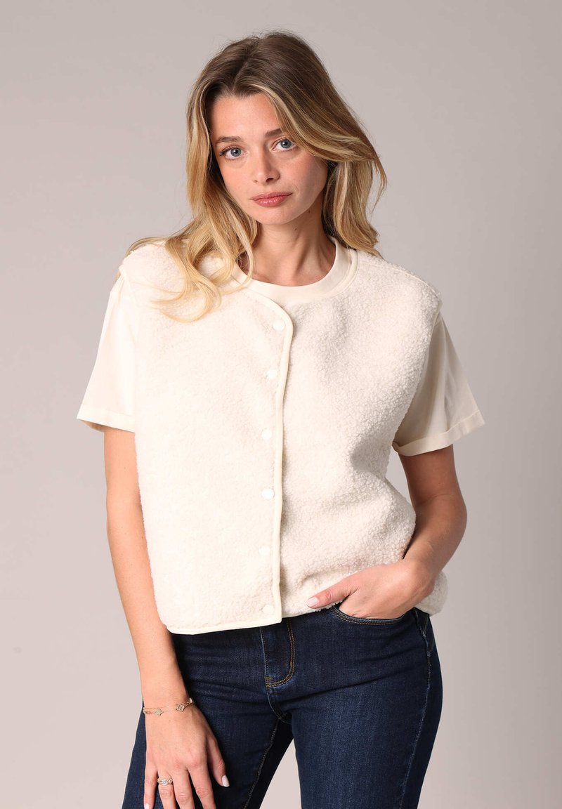 Gilet en polaire blanc avec un col rond, fermeture à pressions à l'avant, et manches courtes porté sur un haut clair, associé à un jean bleu foncé.