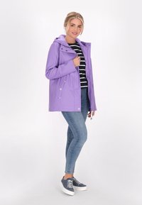 Violetinis nepralaidus vandeniui džemperis su kapišonu, snapų saga ir šoninėmis kišenėmis, dėvimas ant juodai baltos juostuotės marškinėlių ir mėlynų džinsų.