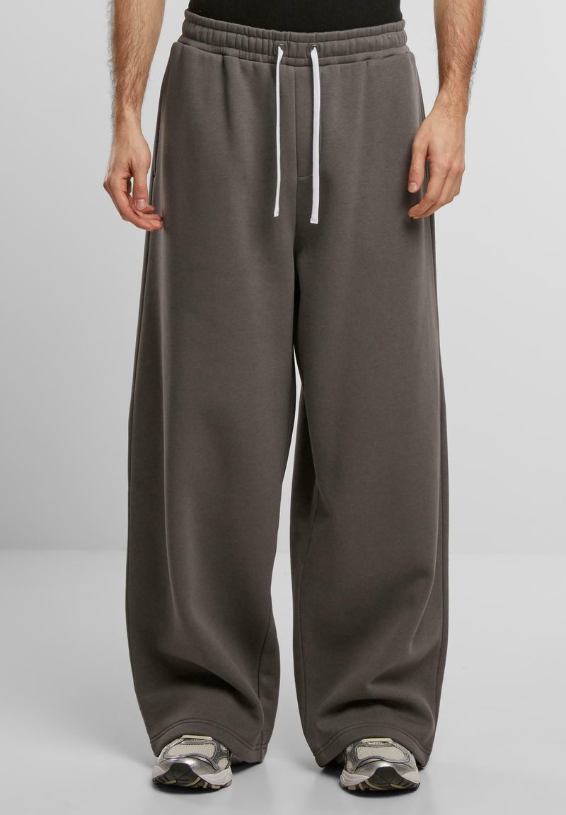 Pantaloni della tuta grigi con una vestibilità ampia e rilassata, realizzati in un tessuto morbido. Presentano una vita con cordino e dettagli bianchi. Indossati con scarpe da corsa.