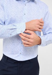 Camicia azzurro chiaro con maniche lunghe, motivo paisley, e bottoni scuri in contrasto. Tessuto morbido e vestibilità su misura.