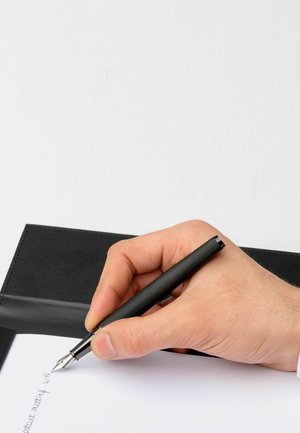 Schwarzer strukturierter Füllfederhalter, der in einer Hand gehalten wird und auf weißem Papier schreibt. Im Hintergrund ist ein schwarzes Ledernotizbuch zu sehen.