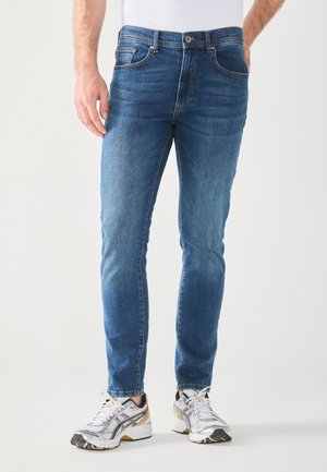 Homme portant un jean bleu slim fit et des baskets blanches, debout devant un fond clair uni.
