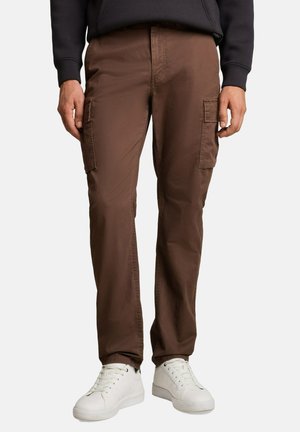 Man draagt bruine cargobroek met zijzakken, witte sneakers en een zwarte longsleeve, staand tegen een neutrale achtergrond.