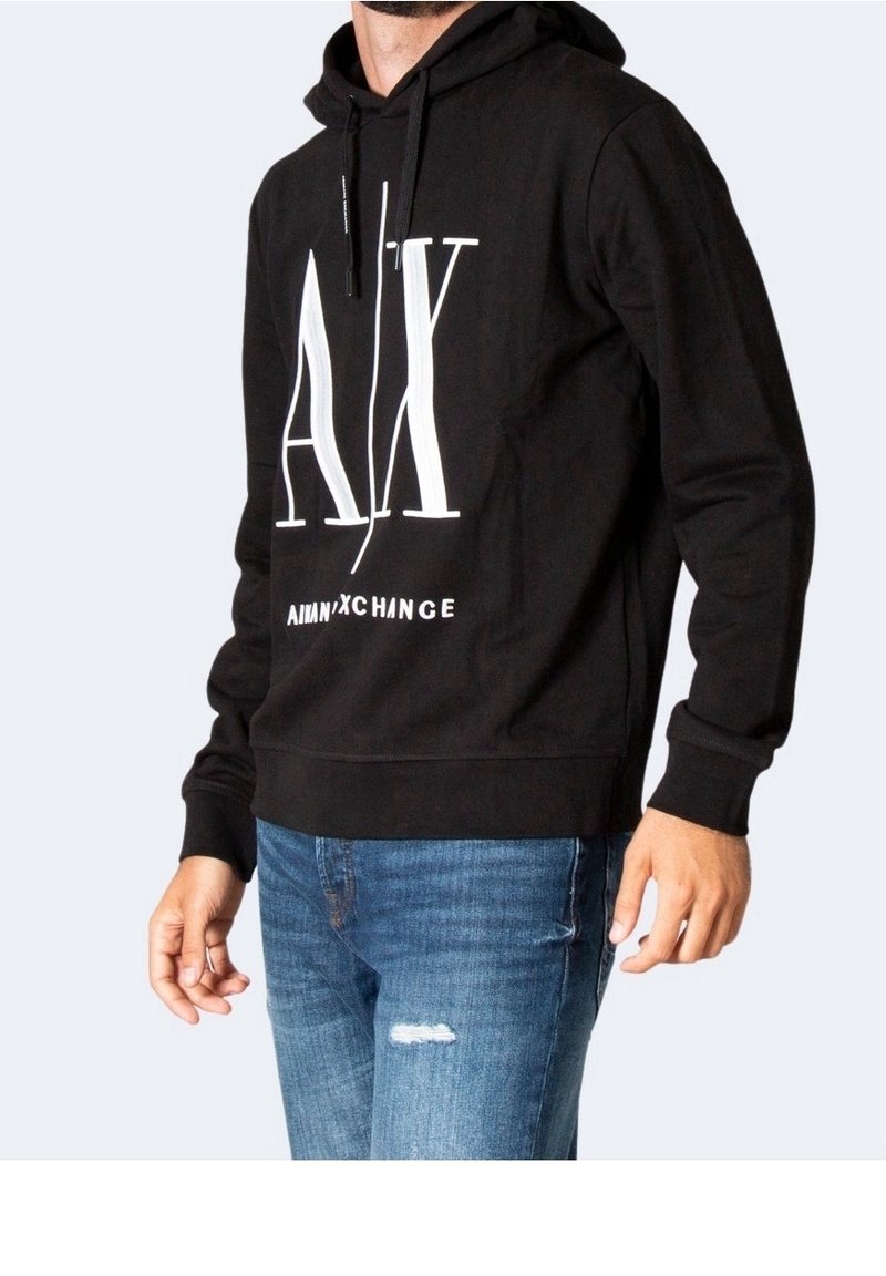 Homme portant un sweat à capuche noir avec un grand logo blanc "A|X" et un jean bleu délavé avec une petite déchirure sur la cuisse gauche.