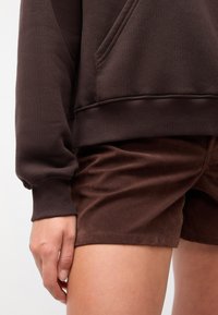 Hanorac maro cu buzunar frontal și manșete ribbed, purtat cu pantaloni scurți maro asortați, având o textură moale.