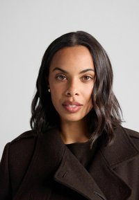 Cappotto di lana marrone con ampi revers e bottoni doppi, indossato sopra un maglione scuro. I capelli sono scuri e leggermente mossi. Sfondo chiaro.