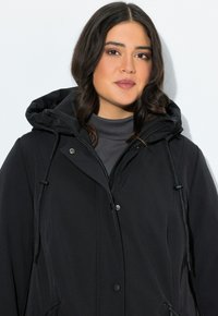 Femme aux cheveux bruns portant une veste à capuche noire avec fermeture éclair et boutons sur un haut gris foncé, se tenant devant un fond blanc.