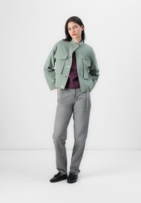 Zielona krótka kurtka z dwiema przednimi kieszeniami, w zestawie z ciemną bluzką i szarymi spodniami o prostym kroju. Czarne buty dopełniają outfit.