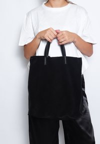 Borsa tote in suede nero con bordi scallopati, manici in pelle e cuciture bianche. Il modello tiene la borsa indossando una t-shirt bianca e pantaloni neri.