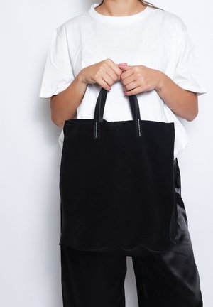 Zwarte suede tote bag met geschulpte randen, leren handvatten en witte stiksel. Model houdt de tas vast en draagt een wit t-shirt en zwarte broek.