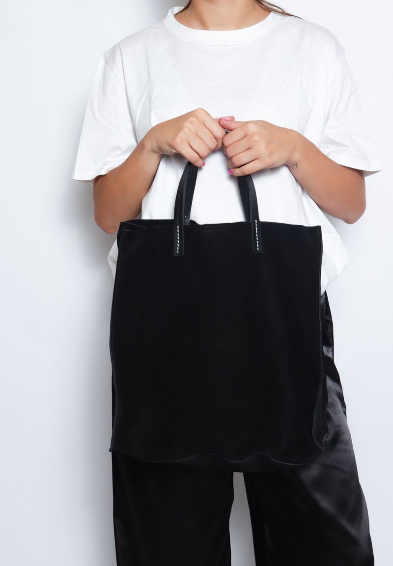 Borsa tote in suede nero con bordi scallopati, manici in pelle e cuciture bianche. Il modello tiene la borsa indossando una t-shirt bianca e pantaloni neri.