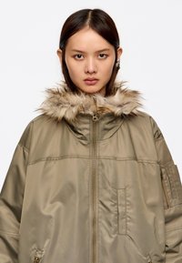 Parka verde oliva con capucha de pelo sintético, cierre frontal de cremallera y bolsillos laterales. Hecha de material suave y ligero.