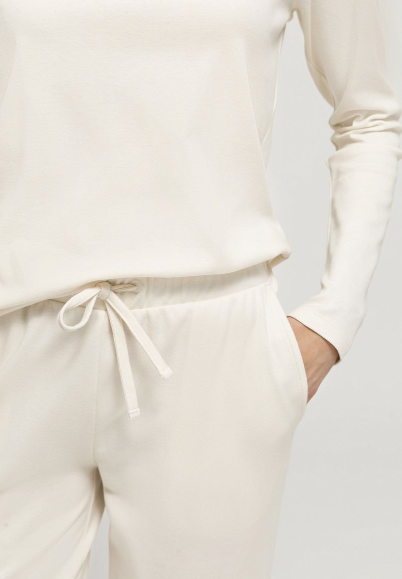 Ljusbeige loungewear-set bestående av en långärmad topp och byxor med elastisk midja och dragsnöre. Mjuk, slät tygtextur.