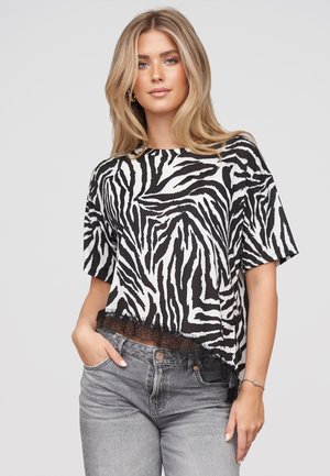 Ung kvinde med blondt hår bærer en sort-hvid zebrastribet kortærmet top med blondekant og grå jeans, poserer med den ene hånd i lommen.