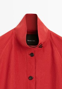 Rode jas met hoge kraag en zwarte knopen, gemaakt van gestructureerde stof, met een Massimo Dutti-label aan de binnenkant van de kraag.