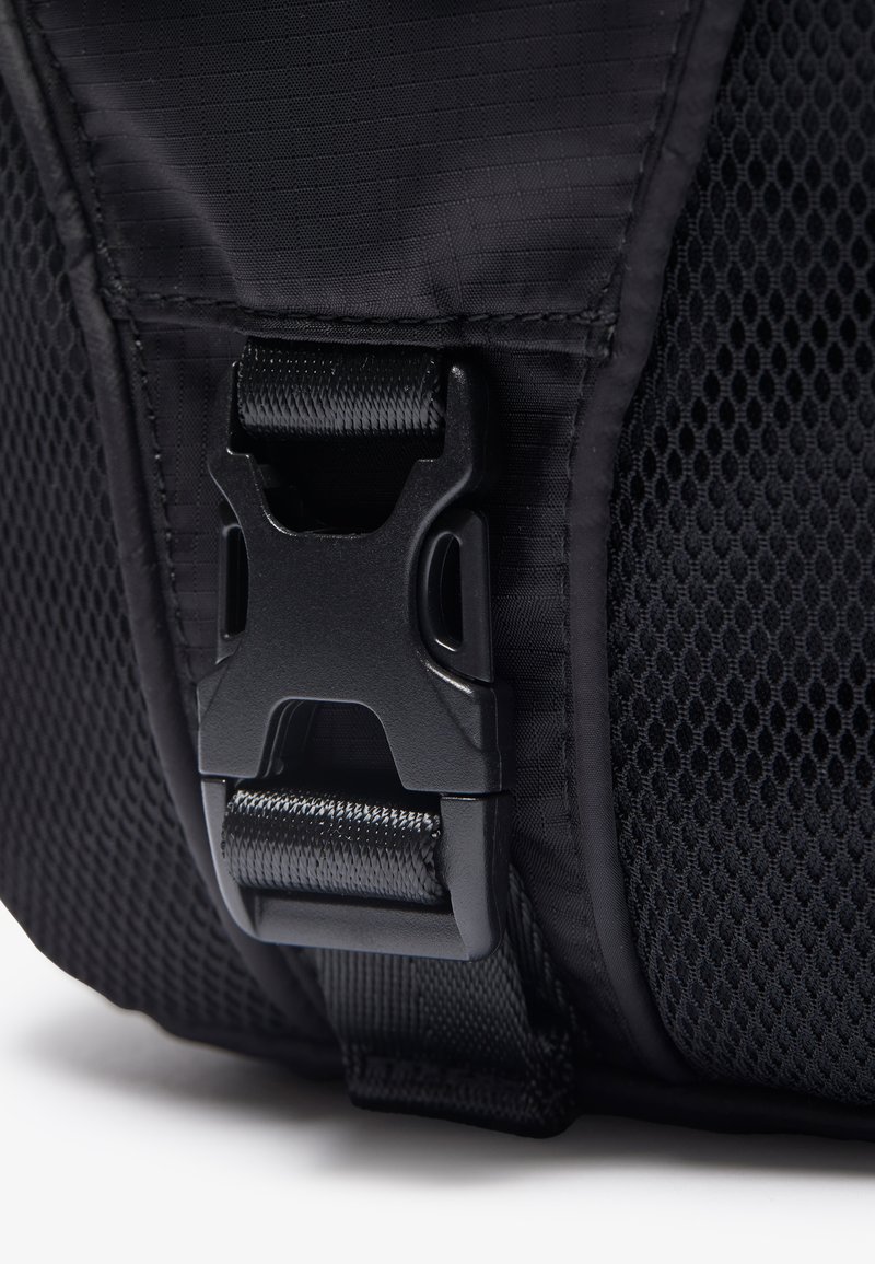 Bolsa de tela negra con cierre de hebilla de plástico. La textura incluye secciones suaves y paneles de malla para ventilación.