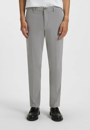 Pantalons gris ajustés en tissu lisse, dotés d'un devant plat, de passants de ceinture et d'une fermeture éclair, portés avec des mocassins noirs.