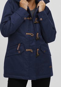 Mörkblå parka med huva, utrustad med knäppningar med toggle, två framfickor och bruna repdetaljer. Tillverkad av strukturerad tyg.