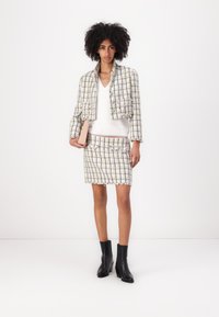 Veste en tweed au design court, motif à carreaux multicolores ; jupe en tweed assortie. Le mannequin tient une pochette beige et porte des bottines noires.